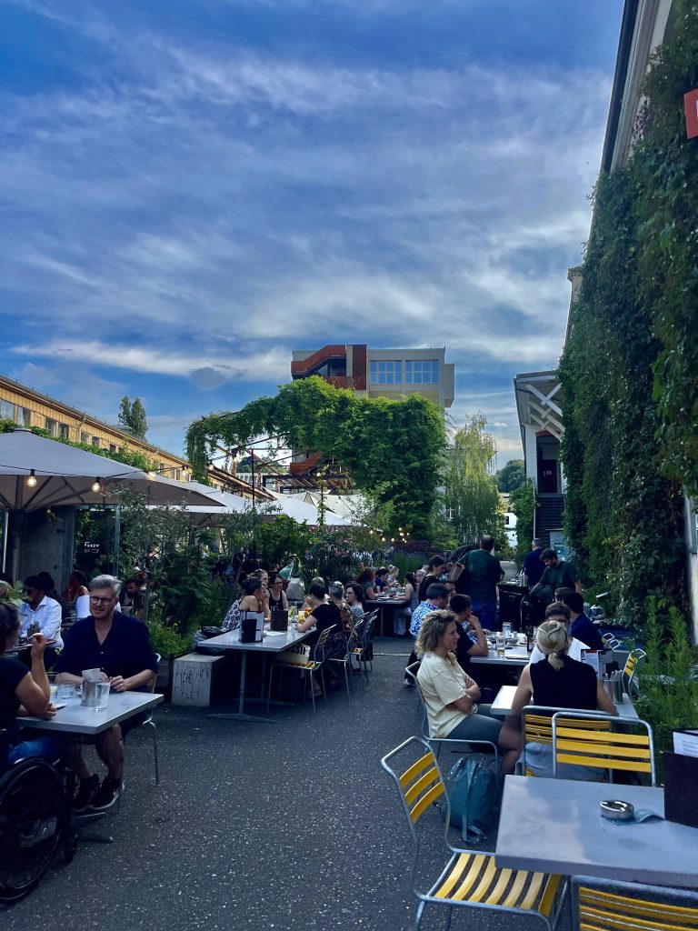 Swiss people eating in outdoor dining terrace surrounded by buildings covered in plants. Showcasing European cultural habits and food traditions.